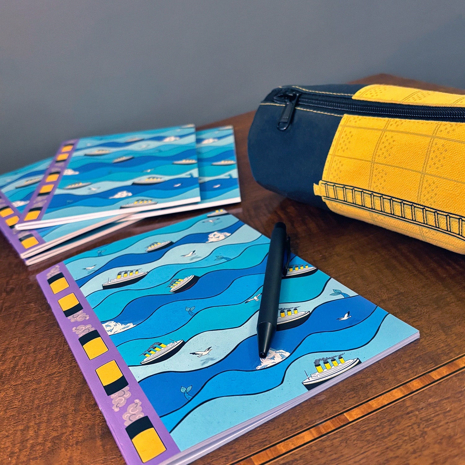 A notebook and pencil case on a table, which were designed by Merpola for Titanic Merch.